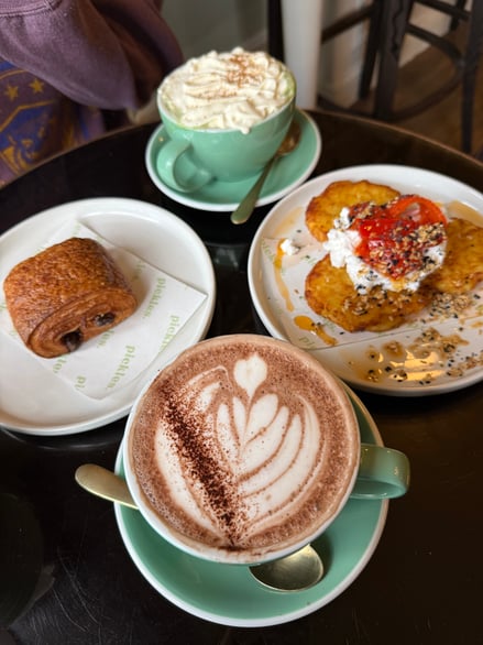 Set in a cafe the image captures a hot chocolate decorated with latte art, as well as some food on two other plates and another drink in the background.
