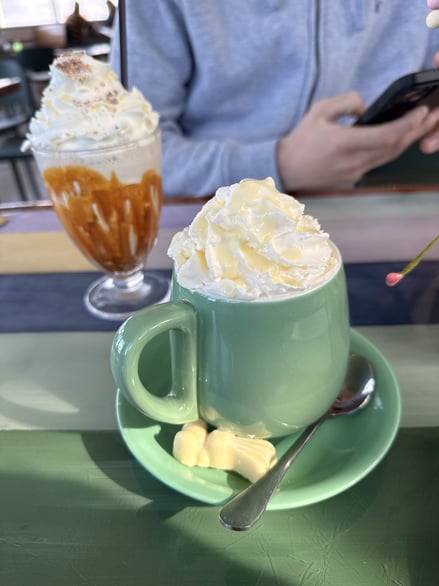 Green mug with whipped cream hot chocolate and caramel dessert in background