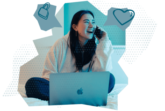 A female student is sat behind a laptop on her bed, talking on her phone with a smile on her face