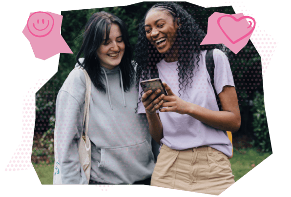 Two smiling female students are looking at a phone screen together, with leafy surroundings in the background