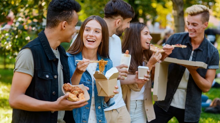 A group of students are stood around eating takeaway meals and chatting outdoors in a park.